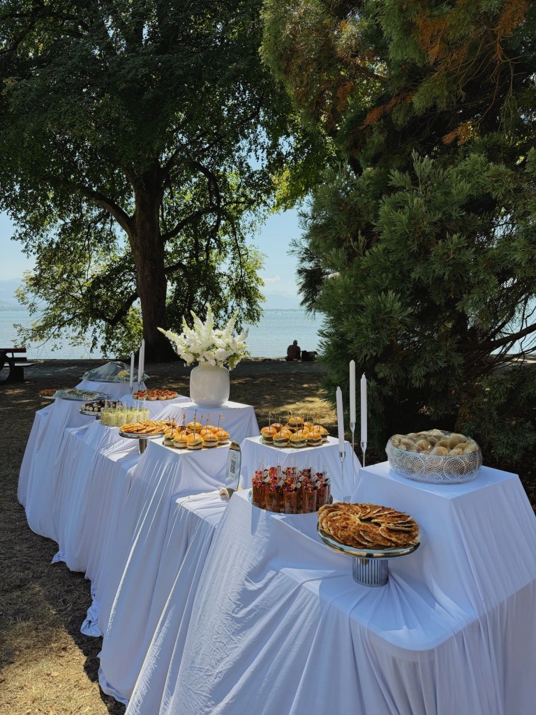 Lange weiße Buffettafel im Freien unter einem Baum, dahinter der See