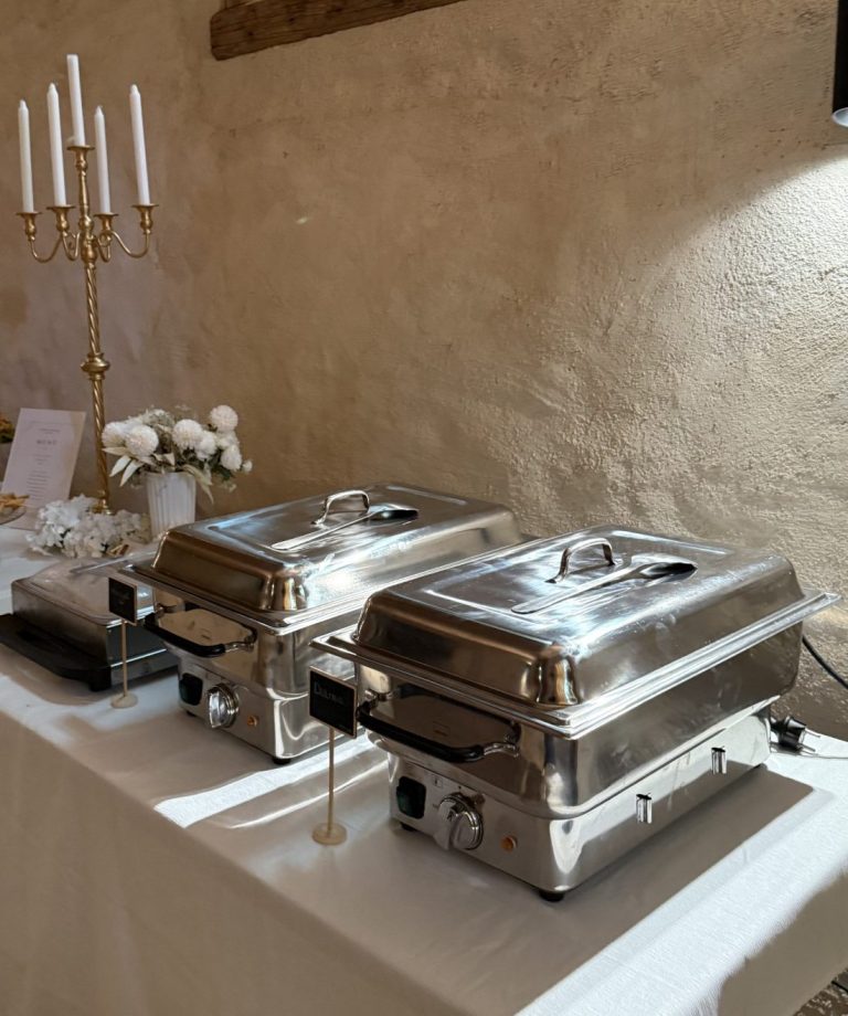 Zwei silberne Warmhaltebehälter auf weißer Tafel, daneben Kerzenleuchter und Blumen
